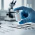 Gloved hand holding a small clear vial next to pipettes and laboratory equipment in a clean research setting.